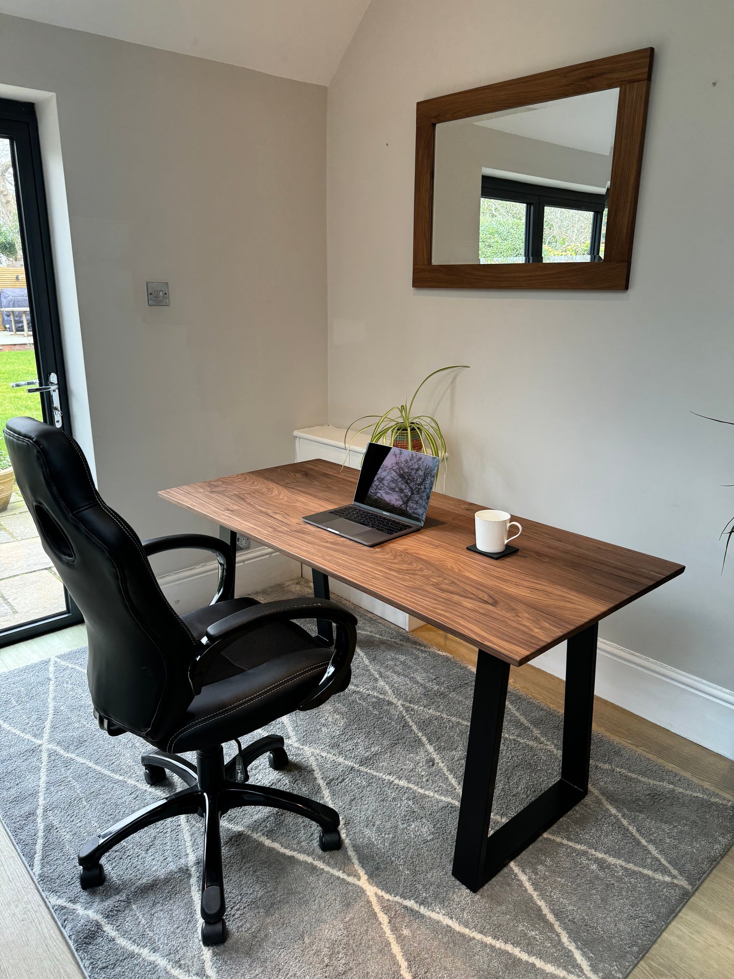 A solid walnut wood office desk with black metal legs, a laptop on the desk, and a cup to the side, placed against a wall with a large mirror above it, and a decorative plant to the side.