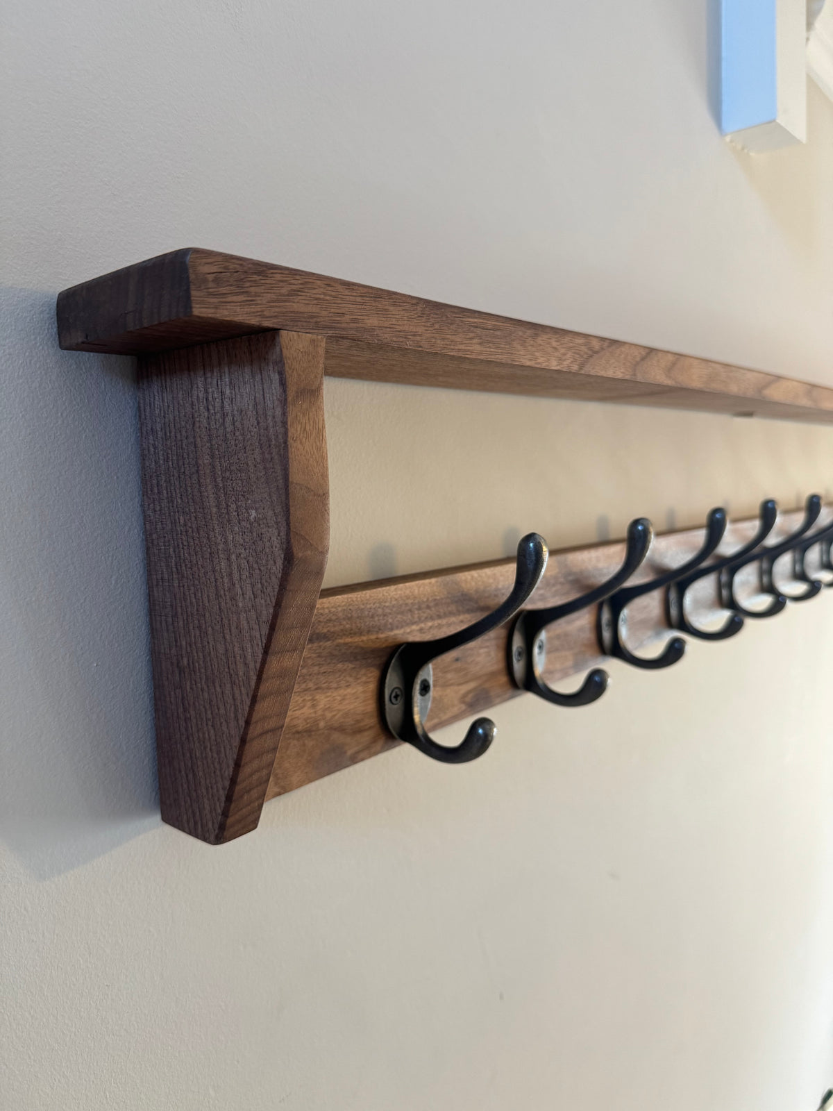 A handmade solid dark American walnut wooden coat rack with a shelf, featuring black cast iron coat hooks against a white wall.