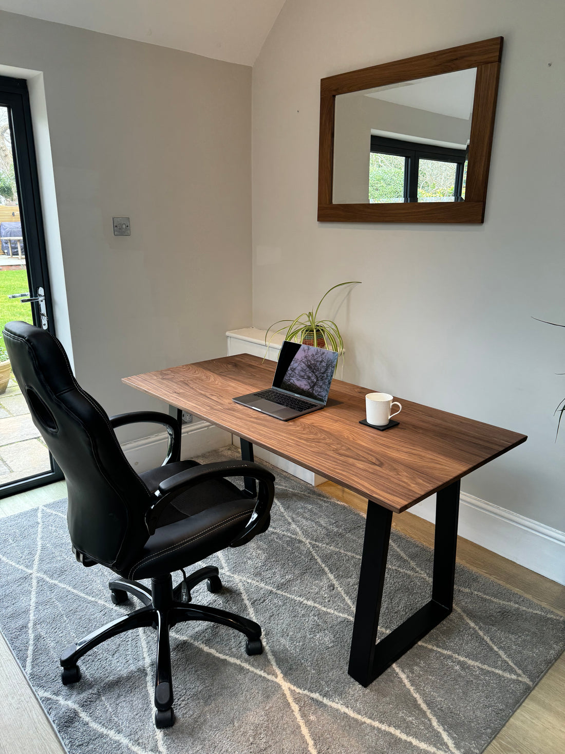 A solid walnut wood office desk with black metal legs, a laptop on the desk, and a cup to the side, placed against a wall with a large mirror above it, and a decorative plant to the side.